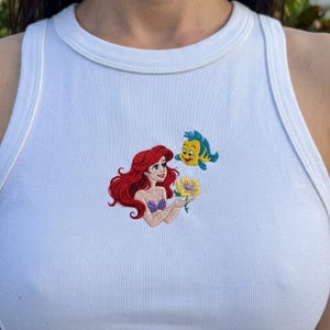 Puede incluir: Camiseta sin mangas blanca con un dise&ntilde;o bordado de Ariel de La Sirenita, sosteniendo una flor amarilla, con Flounder. El dise&ntilde;o tiene colores rojo, azul, amarillo y morado.