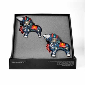 Puede incluir: Dos figuras decorativas de toros azules con diseños florales y de volutas, presentadas en una caja negra con interior gris. Los toros tienen cuernos plateados y están adornados con detalles rojos, amarillos y blancos. La caja dice "PERUVIAN ARTCRAFT".