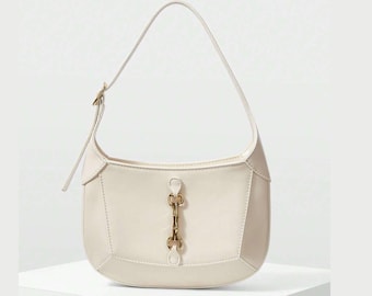 May include: Cream-colored shoulder bag with a curved shape and a single strap. The bag features a gold-colored clasp and stitching details. The bag is sitting on a white surface.