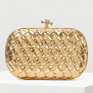 May include: A gold clutch bag with a woven pattern. The rectangular bag has rounded edges and a decorative clasp. The metallic finish gives the bag a luxurious appearance. The clutch is a fashion accessory.