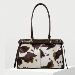 May include: A rectangular handbag with a cowhide pattern in brown and white. The bag has dark brown leather trim, handles, and straps. Gold-coloured buckles add detail. The bag is set against a white background.