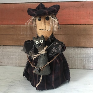 Könnte beinhalten: Handgemachte Halloween-Hexenpuppe mit einem schwarz-burgunderfarbenen karierten Kleid und Hut. Die Puppe hat Knopfaugen und hält eine schwarze Katzenpuppe mit Knopfaugen. Die Hexe hat weißes Haar und einen genähten Mund.