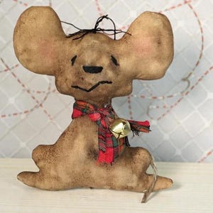 May include: A handcrafted, brown mouse ornament with large ears and a red, green, and black plaid scarf. A small gold bell hangs from the scarf. The mouse has a distressed, vintage appearance, and is likely a holiday decoration.