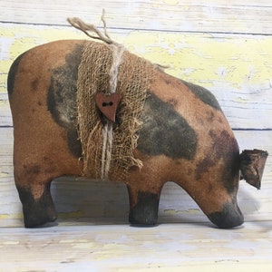 May include: A handmade, rustic pig-shaped decorative item. The pig is primarily brown with dark patches, and is adorned with a burlap-like wrap around its neck, secured by a wooden heart-shaped button. The pig's snout is bent downward.