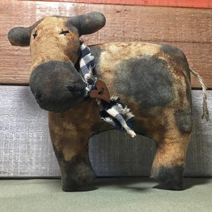 Può includere: Statuetta di mucca di peluche dall'estetica rustica. La mucca è principalmente marrone con macchie grigie e dettagli neri. Indossa una sciarpa a scacchi blu e bianca con un cuore di legno. La mucca ha un viso cucito e una piccola coda.
