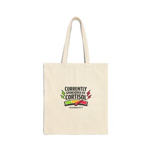 May include: Cream-colored tote bag with the text "CURRENTLY SPONSORED BY CORTISOL" in black, green, red, and white. The bag has a graphic of a stress meter and the words "streaming 24/7" below the text.