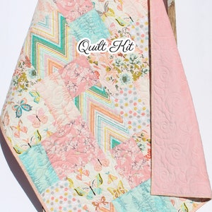 May include: A pink and blue patchwork quilt kit with a floral, butterfly, and chevron pattern. The kit includes fabric for the quilt top, backing, and binding. The text "Quilt Kit" is visible on the quilt top.