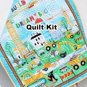 May include: A quilted baby blanket with a construction theme. The quilt features yellow construction vehicles, buildings, and the words "BUILD," "DREAM BIG," and "EXPLORE." The quilt has a blue border and a light blue background with a swirling pattern.