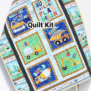 May include: A colorful quilt kit featuring construction vehicles and the words "Build, Explore, Play, Dream." The quilt has a light blue background with square panels showcasing trucks, excavators, and cement mixers. The text "Quilt Kit" is also visible.