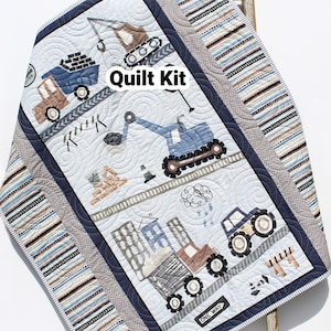 May include: A quilt kit featuring a construction theme with blue and brown trucks, a crane, and a one-way sign. The quilt kit is on a white background with a blue and gray border. The text "Quilt Kit" is written in white on a blue background.