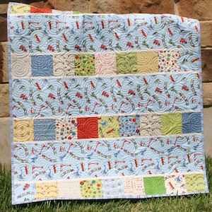 Baby quilt pattern featuring Riley Blake train fabrics with colorful charm pack squares and novelty transportation prints in a modern patchwork nursery quilt design.