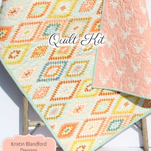 May include: A quilt kit featuring a geometric pattern in shades of orange, yellow, blue, and green. The back of the quilt is a light pink fabric with a white deer print. The text "Quilt Kit" is written in cursive on the front of the quilt. The text "Kristin Blandford Designs" is written in a white frame at the bottom of the image.