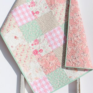 Puede incluir: Una colcha de beb&eacute; de retazos con un borde verde. La colcha est&aacute; hecha de varias telas florales y vichy en tonos de rosa, verde y blanco.
