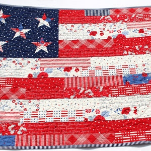 May include: A red, white, and blue patchwork quilt featuring a large American flag design. The flag is made up of different fabric scraps, including stripes, stars, and floral patterns. The quilt is quilted with a simple swirl pattern.