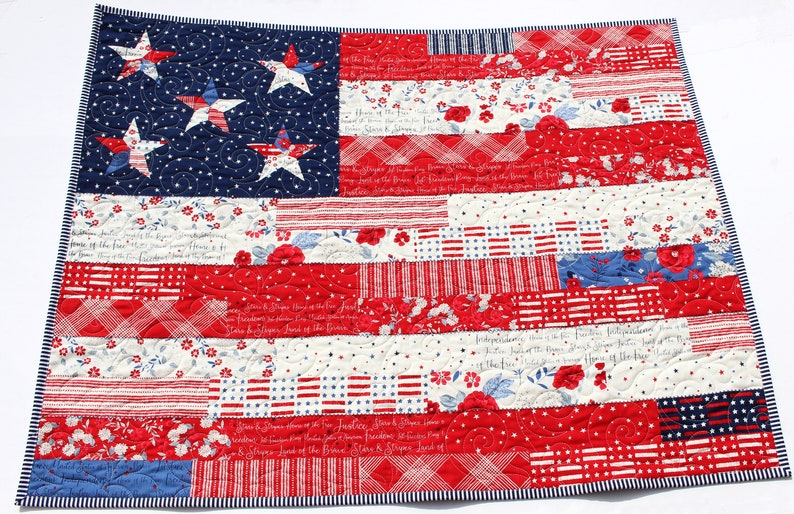 May include: A red, white, and blue patchwork quilt featuring a large American flag design. The flag is made up of different fabric scraps, including stripes, stars, and floral patterns. The quilt is quilted with a simple swirl pattern.