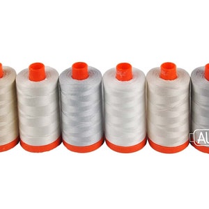 May include: Six spools of thread in various shades of white and gray. The spools are arranged in a row and have orange tops.