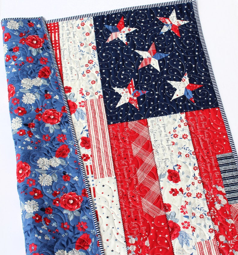 May include: A red, white, and blue patchwork quilt with a floral pattern on the back. The front of the quilt features a traditional American flag design with stars and stripes. The quilt is quilted with a swirling pattern.