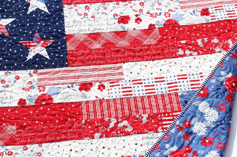 May include: A red, white, and blue patchwork quilt with a patriotic theme. The quilt is made of different fabrics with stars, stripes, and floral patterns. The quilt is quilted with a swirl pattern. The text "Stars & Stripes Home of the Free Justice Freedom for Freedom Land of the Brave Stars & Stripes Land of the Brave" is visible on the quilt.