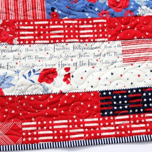 May include: A red, white, and blue patchwork quilt with a patriotic theme. The quilt features a variety of fabrics, including stars, stripes, and floral patterns. The quilt is quilted with a swirling pattern and has a blue and white striped border. The text "Independence Home of the free Freedom Independence Home of the es Justice Land of the Brave Stars & Stripes Justice Land of the Bran &bull; of the Free United States of America Home of the Free United States" is printed on the quilt.