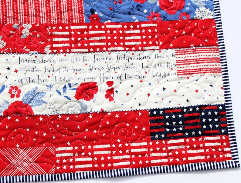 May include: A red, white, and blue patchwork quilt with a patriotic theme. The quilt features a variety of fabrics, including stars, stripes, and floral patterns. The quilt is quilted with a swirling pattern and has a blue and white striped border. The text "Independence Home of the free Freedom Independence Home of the es Justice Land of the Brave Stars & Stripes Justice Land of the Bran &bull; of the Free United States of America Home of the Free United States" is printed on the quilt.