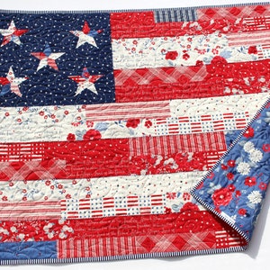 May include: A red, white, and blue patchwork quilt with a design of the American flag. The quilt is made of various fabrics with different patterns, including stars, stripes, and floral prints. The quilt is folded in half and the edges are trimmed with a white and blue striped fabric.