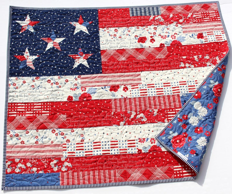 May include: A red, white, and blue patchwork quilt with a design of the American flag. The quilt is made of various fabrics with different patterns, including stars, stripes, and floral prints. The quilt is folded in half and the edges are trimmed with a white and blue striped fabric.