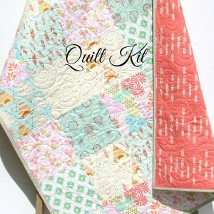 May include: A quilt kit featuring a patchwork design with various floral and geometric patterns in shades of pink, blue, and white. The quilt is edged with a light gray trim and the backing is a solid coral color. The text "Quilt Kit" is written in cursive on the quilt.