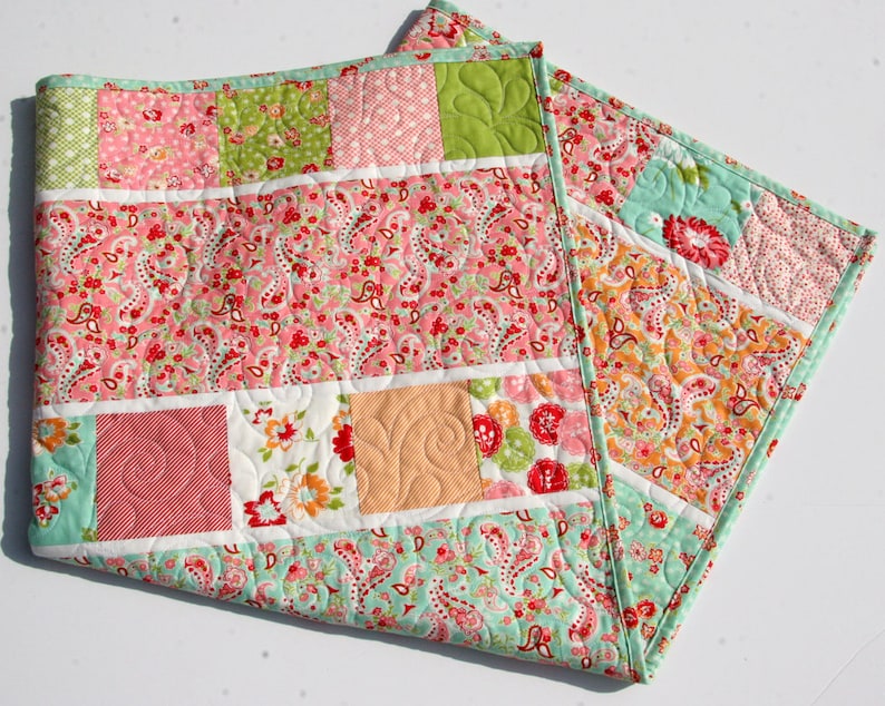 A patchwork quilt with a variety of floral patterns in pink, green, and white. The quilt is made of squares and rectangles, and the edges are finished with a light blue binding.