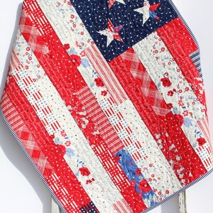 May include: A red, white, and blue patchwork quilt with a star-spangled design. The quilt is made of various fabrics with different patterns, including stripes, stars, and floral prints. The quilt is folded and draped over a wooden ladder.