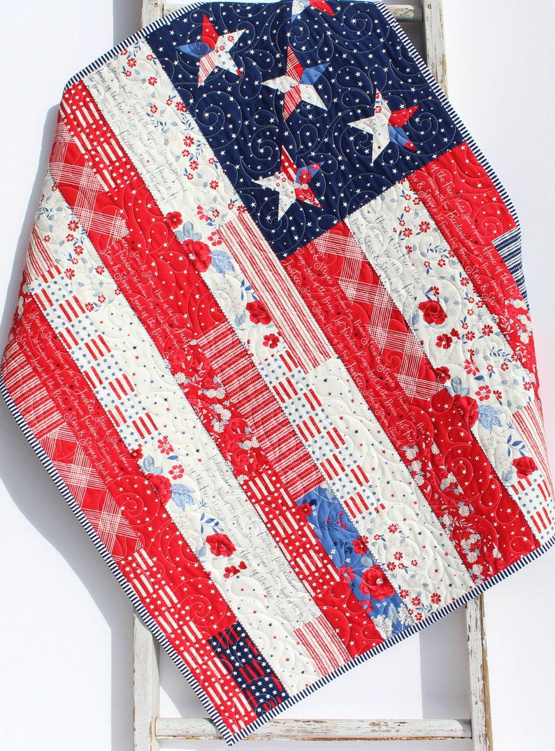 May include: A red, white, and blue patchwork quilt with a star-spangled design. The quilt is made of various fabrics with different patterns, including stripes, stars, and floral prints. The quilt is folded and draped over a wooden ladder.