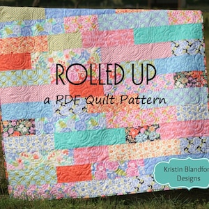 May include: A colorful quilt pattern called "Rolled Up" by Kristin Blandford Designs. The quilt is made of various floral and geometric fabrics in shades of pink, blue, green, and orange.