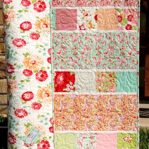 A patchwork quilt with a floral pattern on the back and a colorful patchwork design on the front. The quilt is made with various fabrics in shades of pink, green, orange, and red. The quilt is hanging on a clothesline.
