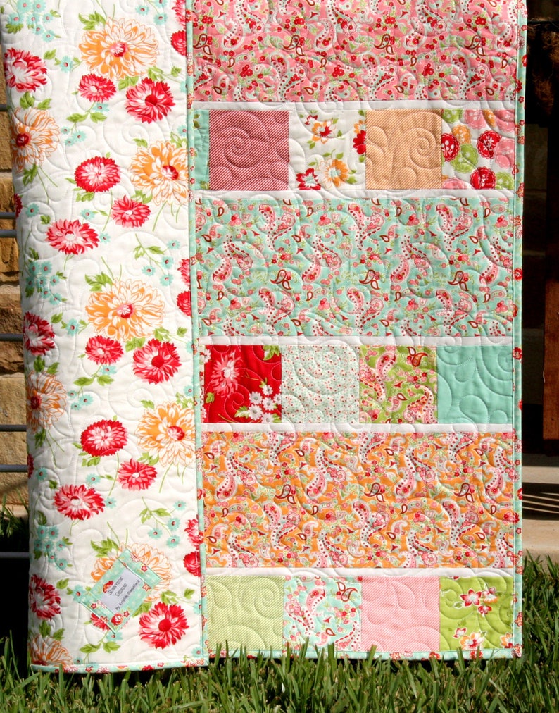 A patchwork quilt with a floral pattern on the back and a colorful patchwork design on the front. The quilt is made with various fabrics in shades of pink, green, orange, and red. The quilt is hanging on a clothesline.