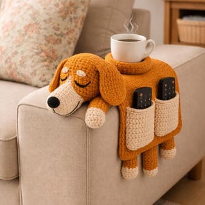 May include: A handmade, orange crochet dog-shaped sofa armrest organizer. It features two pockets for remote controls, a space for a cup, and is resting on a beige sofa. The dog has closed eyes and cream-colored accents.