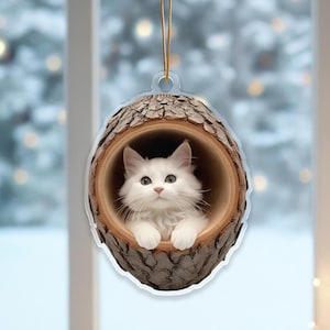 Peut inclure: Ornement décoratif représentant un chat blanc et duveteux à l'intérieur d'un cadre en forme de bûche de bois. Le chat a les yeux verts et regarde vers le haut. L'ornement est suspendu par une ficelle dorée.