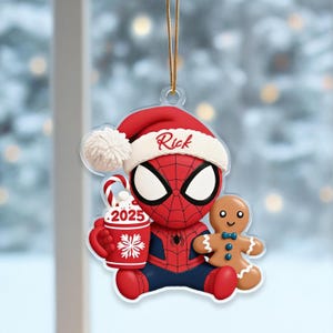 Pode incluir: Enfeite de Natal do Homem-Aranha. O super-herói vermelho e azul usa um gorro de Papai Noel com o nome "Rick". Ele segura uma caneca com "2025" e um desenho de floco de neve, e um boneco de gengibre. Uma corda dourada está presa para pendurar.