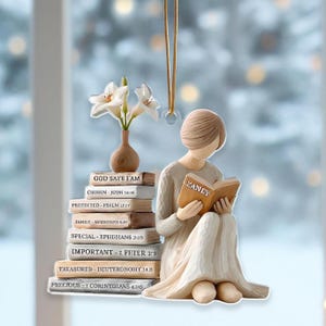Peut inclure: Ornement décoratif représentant une figurine lisant un livre intitulé "LANEY". Une pile de livres avec des phrases religieuses est à côté d'elle. Un petit vase avec des fleurs blanches est également présent. L'ornement est suspendu par une ficelle dorée.