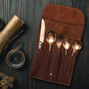 Puede incluir: Un estuche de cubiertos de cuero marrón con un cuchillo, dos cucharas y dos tenedores. Los utensilios tienen un acabado de cobre martillado. El rollo está abierto, revelando la cubertería. Una brújula y un pergamino enrollado están en el fondo.