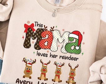 May include: A sand-colored sweatshirt with the text "This Mama loves her reindeer" in a festive design. The word "Mama" is decorated with a red bow and a Santa hat. Below are four reindeer with names: Avery, Leo, Mia, and Ava.