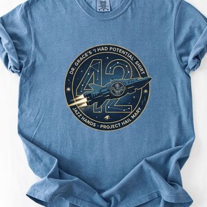 Puede incluir: Camiseta azul vaquero con un diseño gráfico circular que presenta una nave espacial estilizada y el número 42. El texto alrededor del gráfico dice "DR. GRACE'S 'I HAD POTENTIAL SHIRT" y "JAZZ HANDS - PROJECT HAIL MARY."