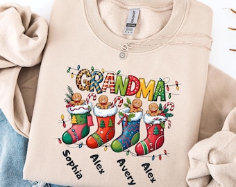 May include: A sand-colored sweatshirt with a Christmas-themed graphic. The graphic features the word "GRANDMA" with a leopard print design, and four Christmas stockings with gingerbread men and names below. A necklace is visible.
