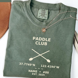 Puede incluir: Una camiseta verde musgo con las palabras "PADDLE CLUB" y remos cruzados impresos en blanco. Debajo, coordenadas, un nombre y "EST. 2025". Un collar dorado con un colgante está sobre la camiseta.