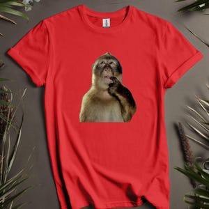 May include: Red t-shirt featuring a graphic of a monkey with its hand near its mouth, as if deep in thought. The monkey has brown and tan fur. The shirt is made of a soft material.