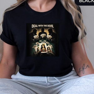 Puede incluir: Camiseta negra con un gráfico temático de póquer. El gráfico muestra tres figuras silueteadas jugando a las cartas, con el texto "DEAL WITH THE DEVIL". La imagen central es una reina en una carta.