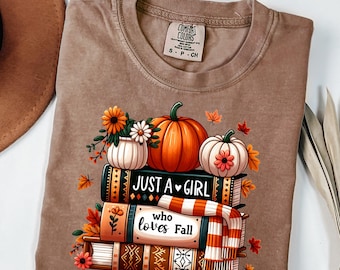 Puede incluir: Camiseta Comfort Colors marrón con un diseño otoñal. El diseño incluye calabazas, flores y libros con el texto "Just a Girl who loves Fall". La camiseta es de color neutro, perfecta para el otoño.