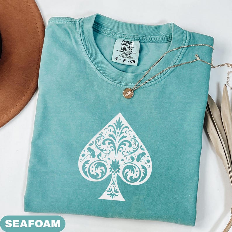 Puede incluir: Camiseta Comfort Colors verde espuma de mar con un dise&ntilde;o de pica blanco. La camiseta tiene cuello redondo y mangas cortas. Un collar de oro con un peque&ntilde;o colgante descansa sobre la camiseta. La camiseta est&aacute; etiquetada S-P-CH.