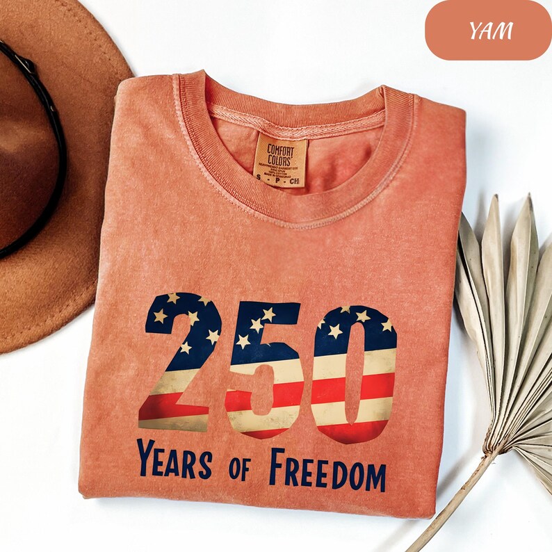 May include: A peach-coloured t-shirt with the text "250 Years of Freedom" in a patriotic design. The numbers are filled with the American flag pattern. The shirt is a Comfort Colors brand.