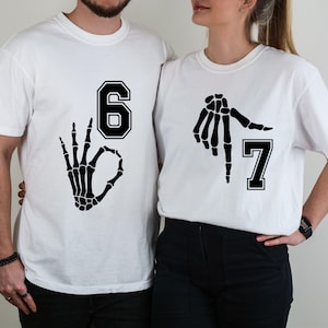May include: Two white t-shirts with black graphics. One shirt features the number 6 with a skeleton hand making an "okay" gesture. The other shirt has the number 7 and a skeleton hand pointing. The shirts are likely for a couple or group.
