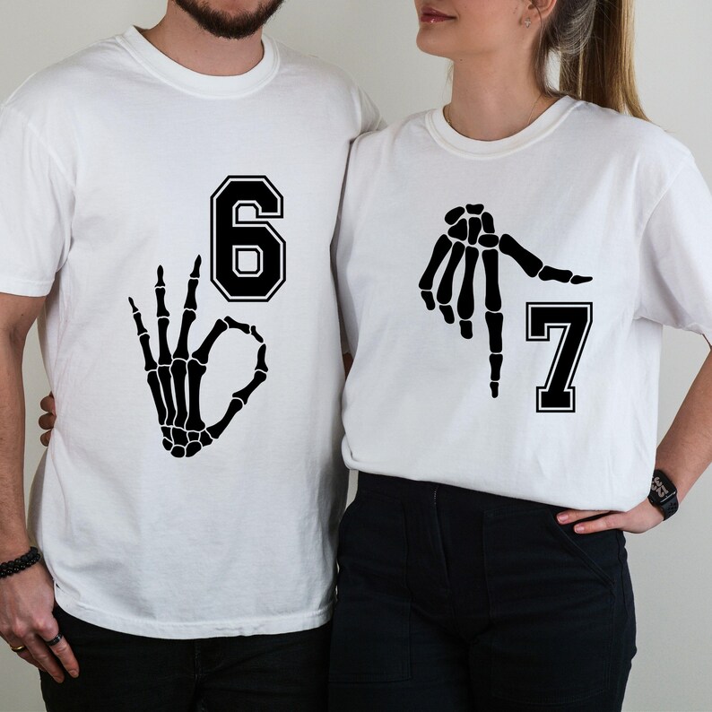 May include: Two white t-shirts with black graphics. One shirt features the number 6 with a skeleton hand making an "okay" gesture. The other shirt has the number 7 and a skeleton hand pointing. The shirts are likely for a couple or group.