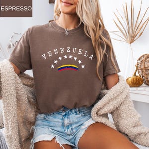 Op de afbeelding: Bruin t-shirt met het woord "VENEZUELA" in witte letters boven een gestileerde Venezolaanse vlag. Het ontwerp bevat witte sterren. Het shirt heeft korte mouwen en lijkt van een zacht materiaal te zijn.